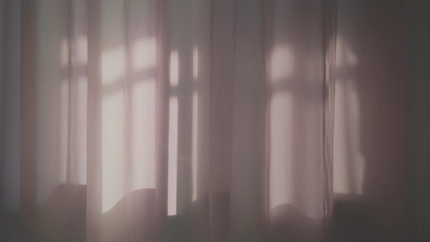 Sunlight casts window frame shadows onto sheer white curtains.